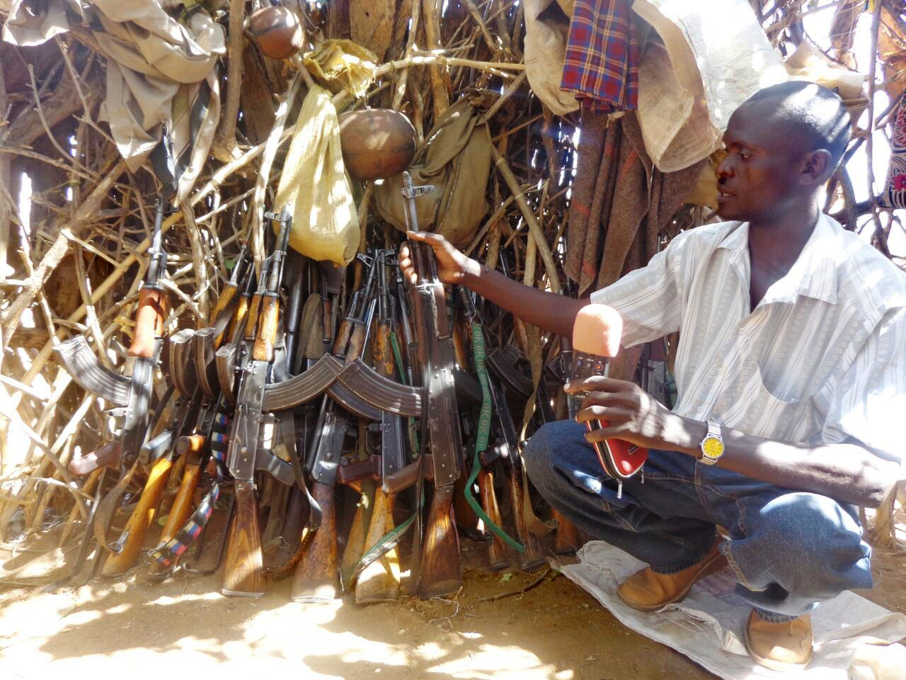 A man sit next to rifles leaning against a bush.